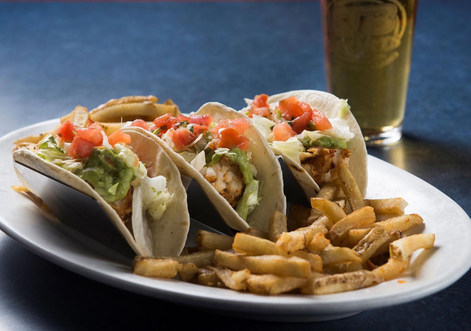 Tacos on a plate with fries, lettuce, salsa, and a drink in the background