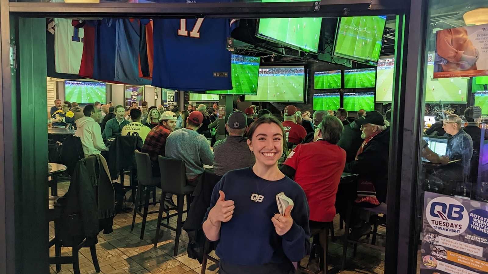 Smiling person giving two thumbs up in a crowded sports bar with many TV screens showing games