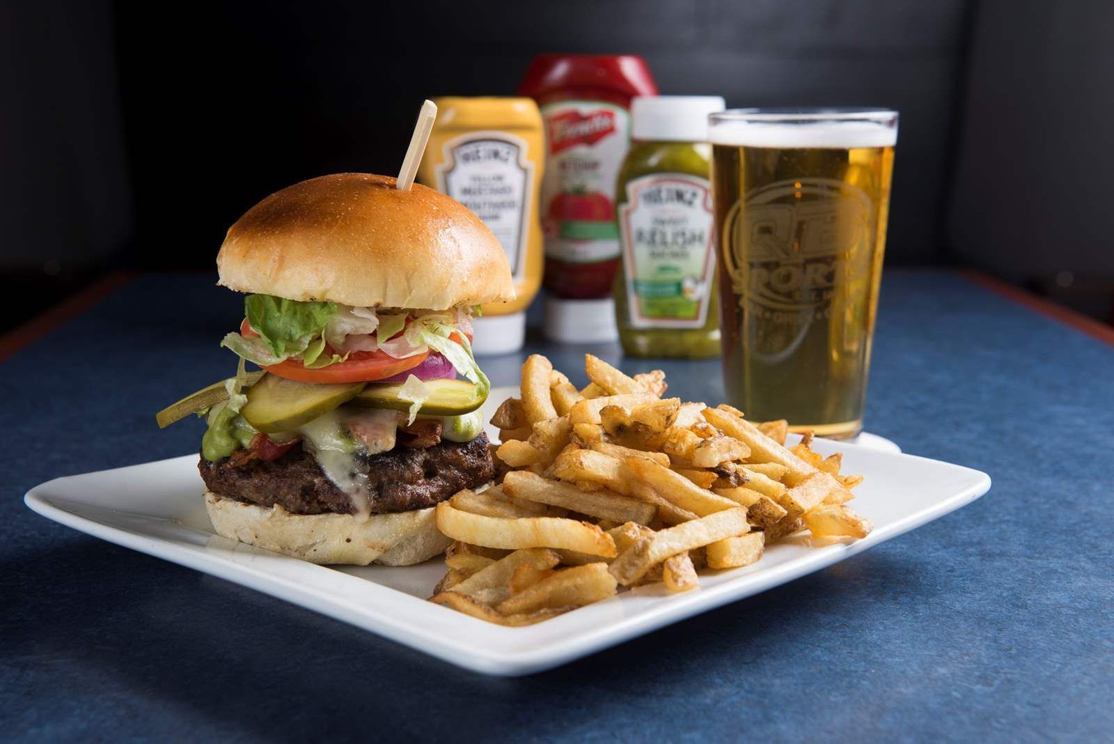Burger and fries on a white plate with ketchup, mustard, and a glass of beer in the background