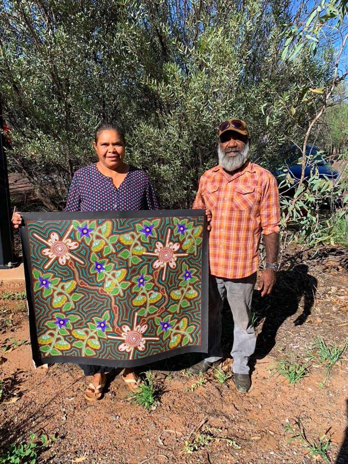 Beaded artwork of a flowering vine with green leaves, brown branches, and pink and blue flowers on a dark background.