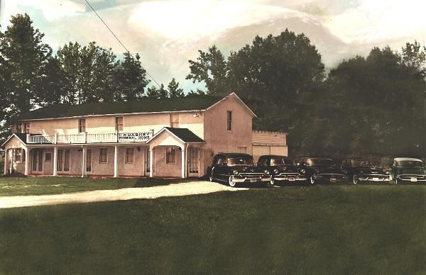 A vintage color photograph of a two-story light-colored building with a long porch, parked next to several black cars.