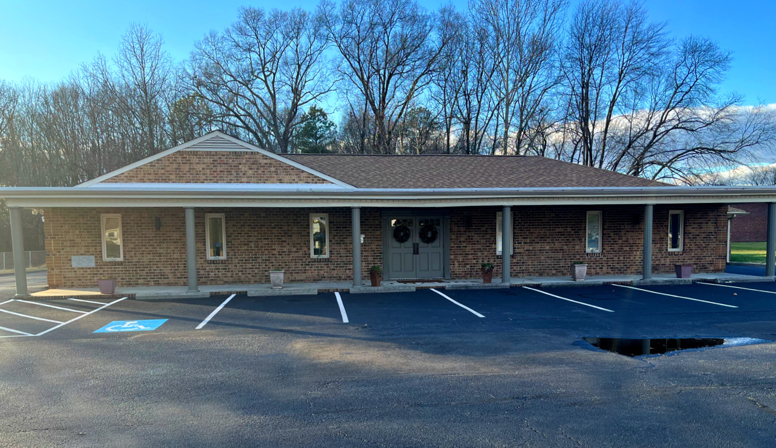 A one-story brick building with a covered porch, front entrance, and a paved parking lot under a bright blue sky.