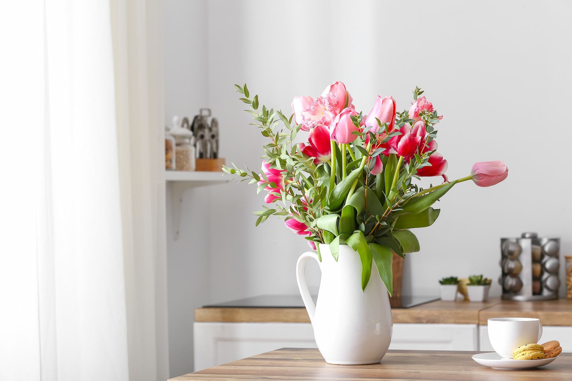 A white pitcher filled with pink tulips and greenery sits on a wooden table next to a cup of coffee.