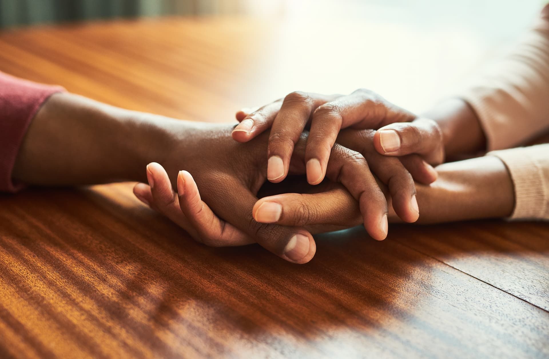 Two pairs of hands resting on a wooden table, with one pair gently holding the other in a gesture of comfort.