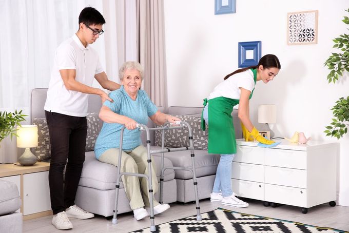 A Man is Helping an Elderly Woman With a Walker in a Living Room — Home Nursing Services In Port Macquarie, NSW