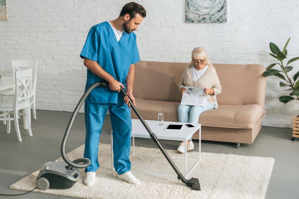 Cleaner Vacuuming a Living Room While a Woman Sits on a Beige Sofa — Home Nursing Services in Parramatta, NSW