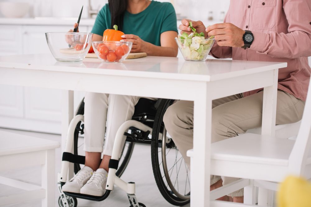 A Woman in a Wheelchair is Sitting at a Table With a Man — Home Nursing Services in Port Macquarie, NSW