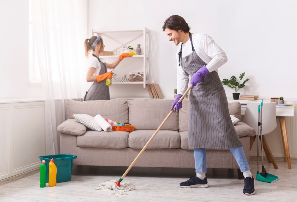 Two People Cleaning a Living Room, One Mopping the Floor Near a Sofa and Shelf — Home Nursing Services in Bankstown, NSW
