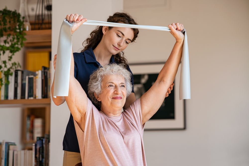 A Woman Is Helping an Elderly Woman with A Resistance Band — Home Nursing Services in Laurieton, NSW