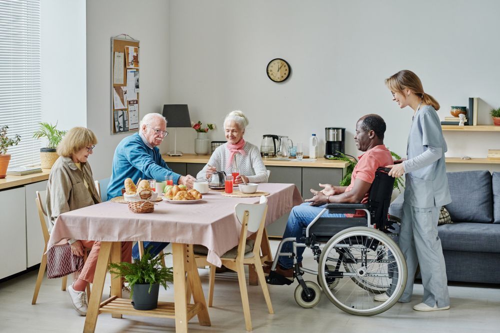 A Group of People Are Sitting at A Table with A Man in A Wheelchair — Home Nursing Services in Laurieton, NSW