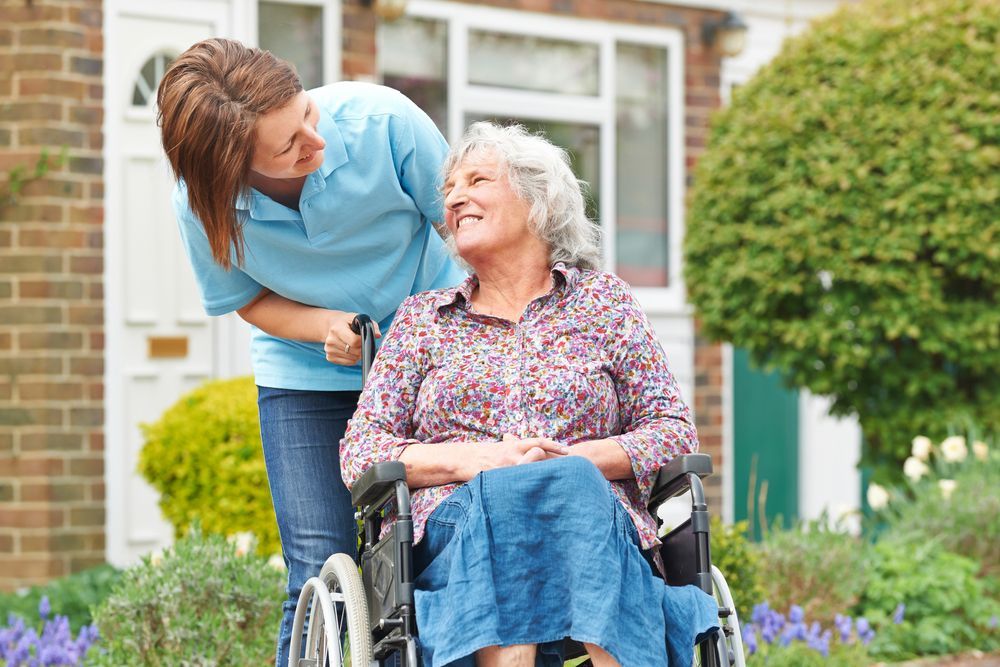 Caregiver Smiles Beside Woman in Wheelchair Outdoors — Home Nursing Services in Liverpool, NSW
