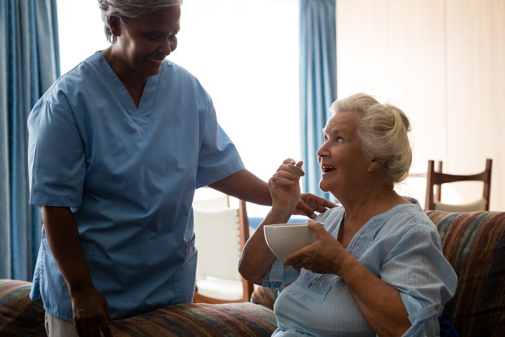Caregiver in Blue Scrubs Serving a Smiling Patient Seated in Bed — Home Nursing Services in Hurstville, NSW