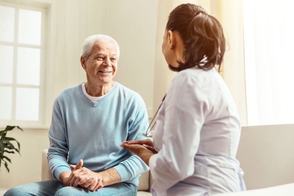 An Elderly Man is Sitting on a Couch Talking to a Doctor — Home Nursing Services in Port Macquarie, NSW