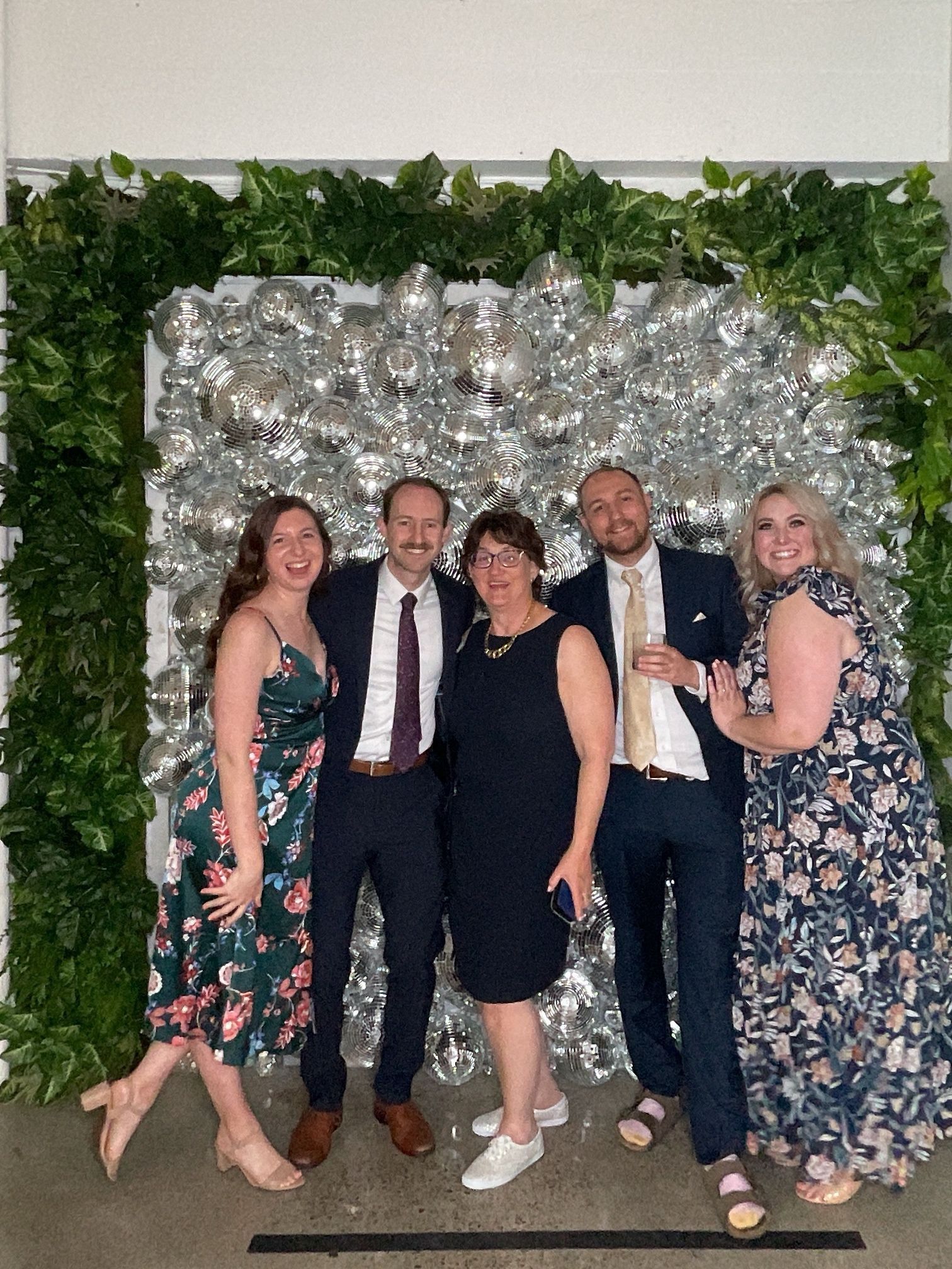 Five people posing and smiling in front of a silver balloon wall and greenery backdrop.