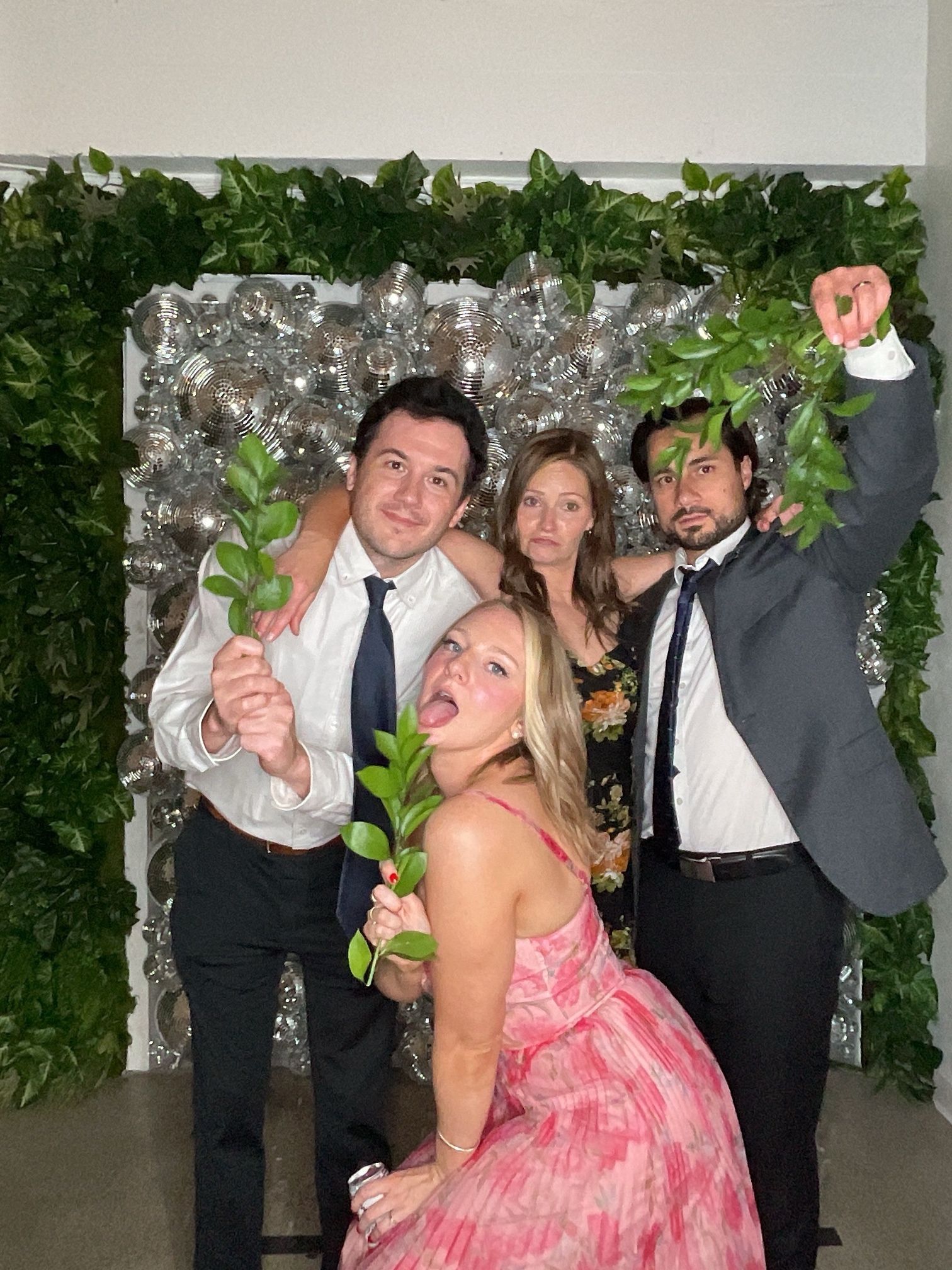 Four people posing at a party backdrop with silver balloons and green foliage, smiling and holding flowers.
