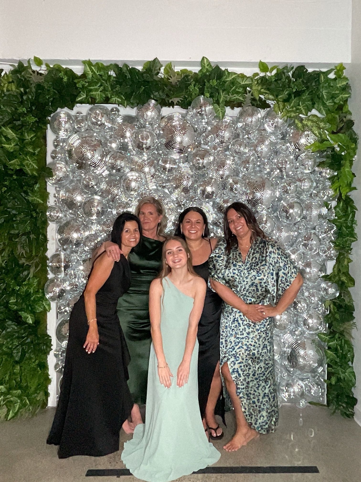 Five people posing and smiling in front of a silver floral backdrop with green leaves at an event photo booth.