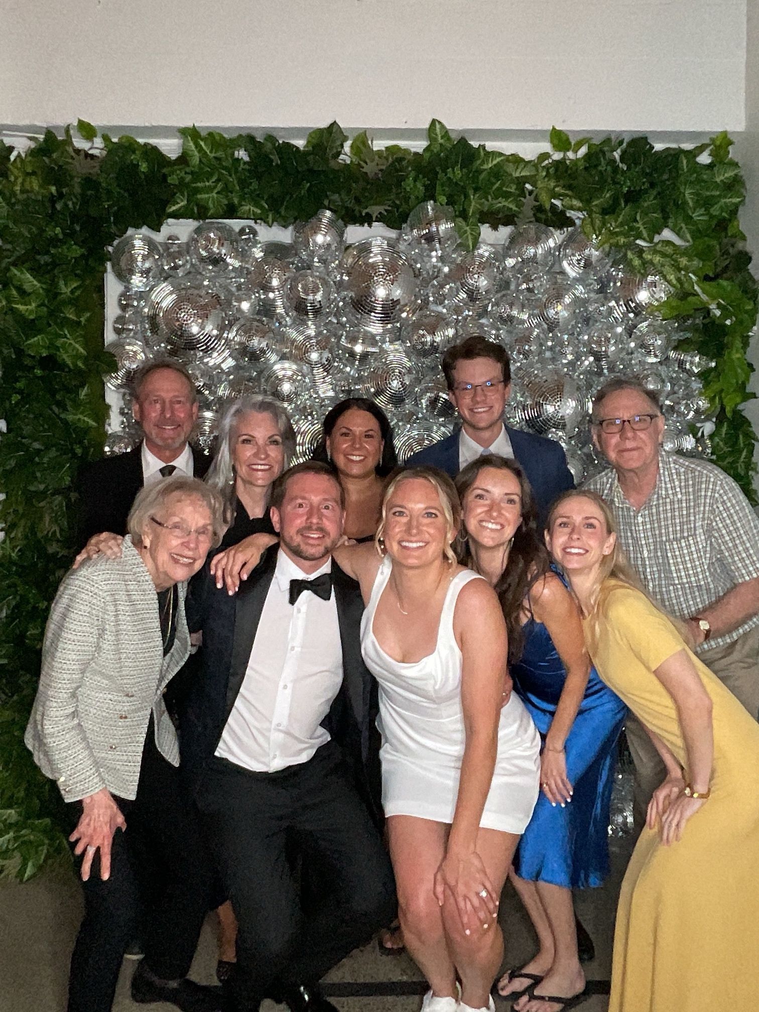 Group of people posing and smiling at a formal event in front of a silver backdrop and greenery.