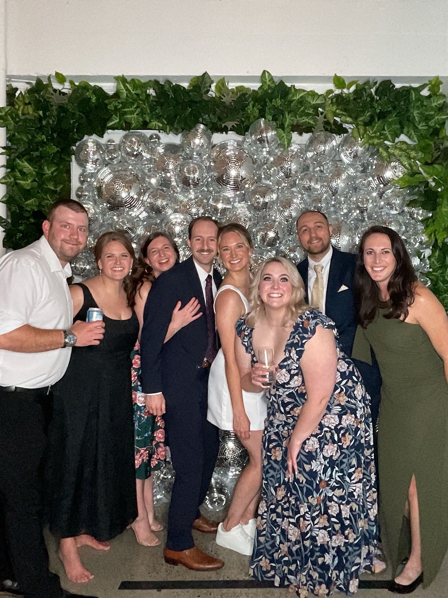 Group of six people posing at an event in front of a silver balloon backdrop and greenery.