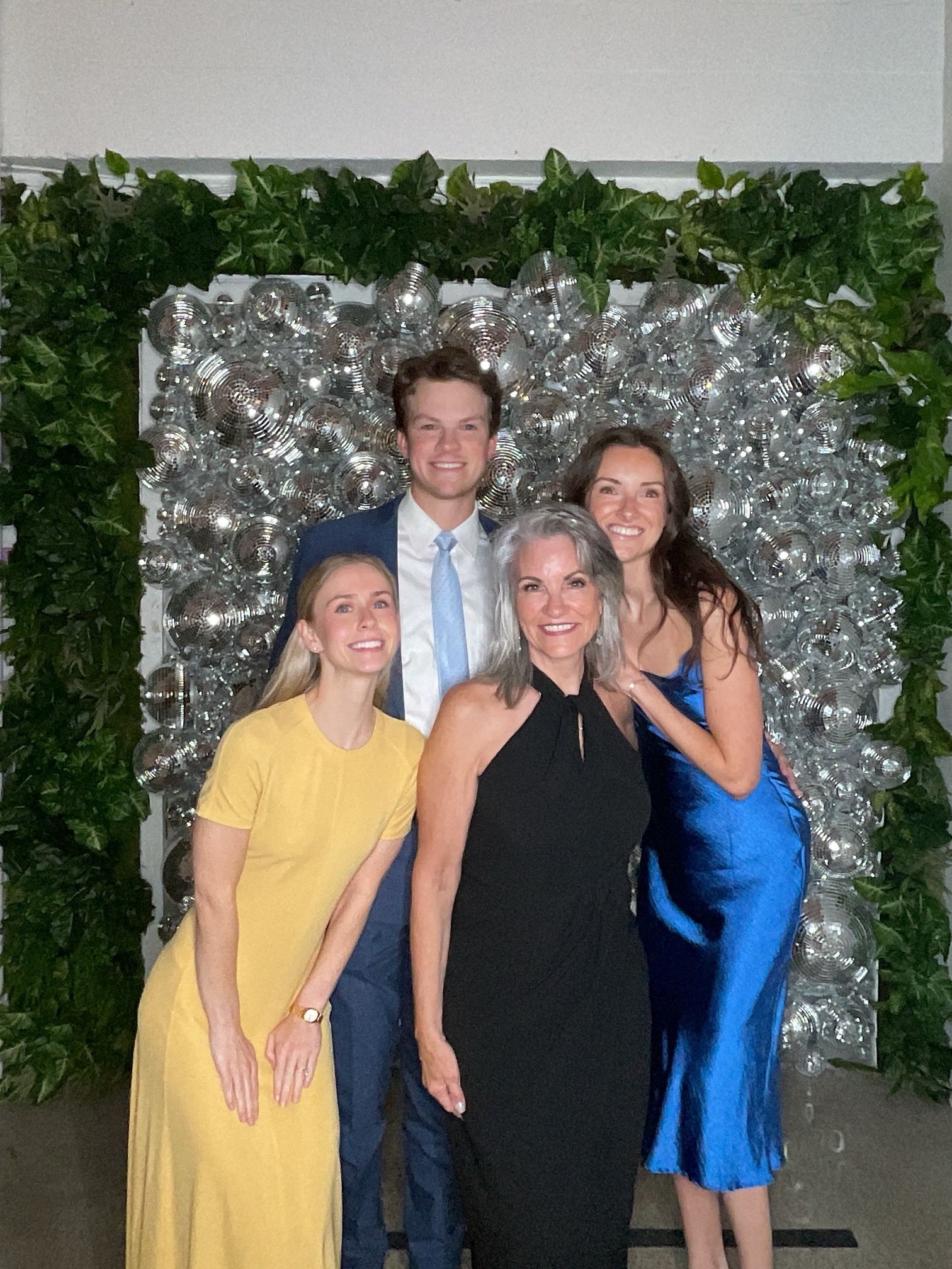 Four people posing in front of a silver floral wall, with green foliage and event lighting.