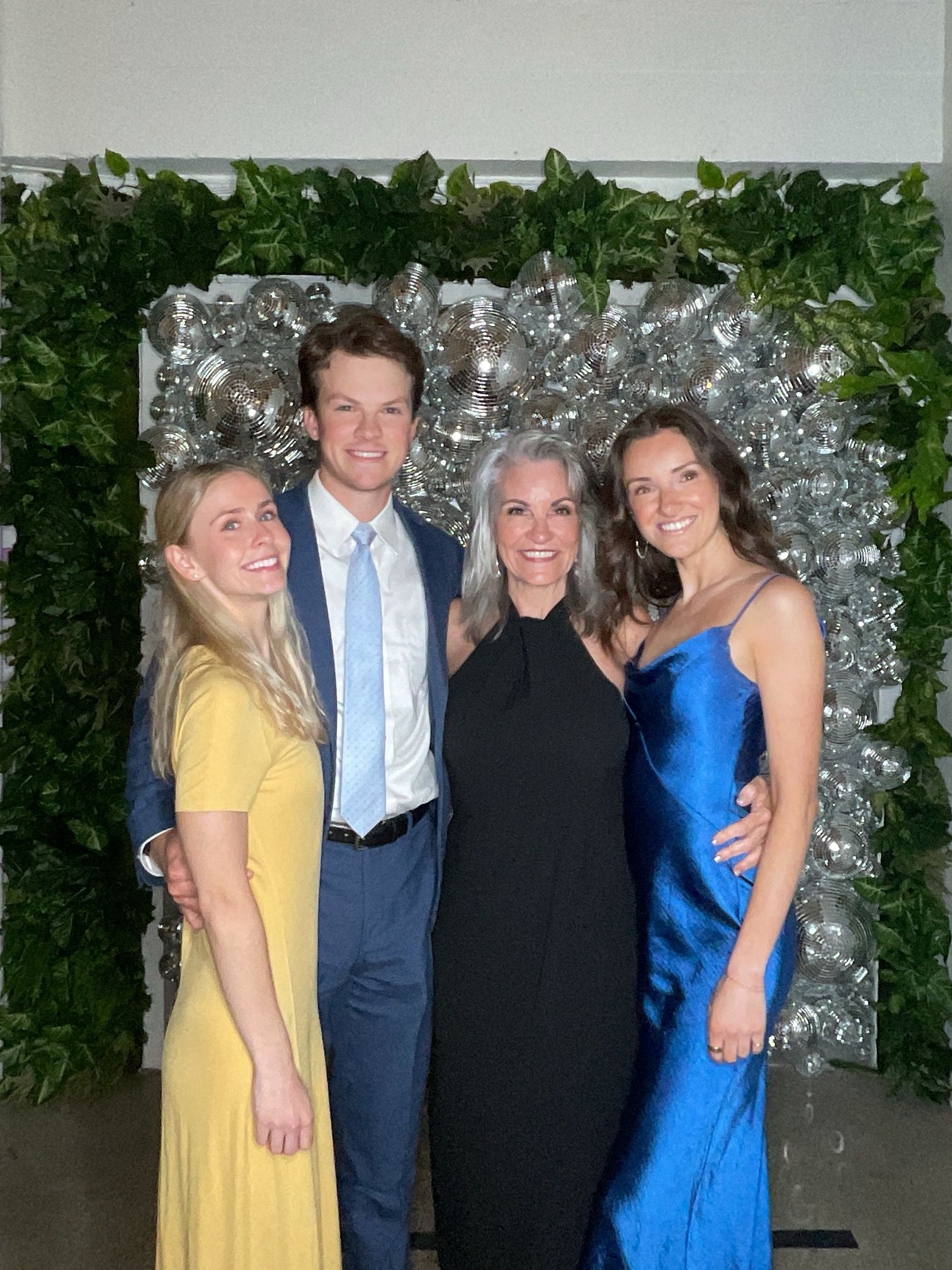 Four people posing together in front of a silver backdrop and greenery wall, dressed in formal attire