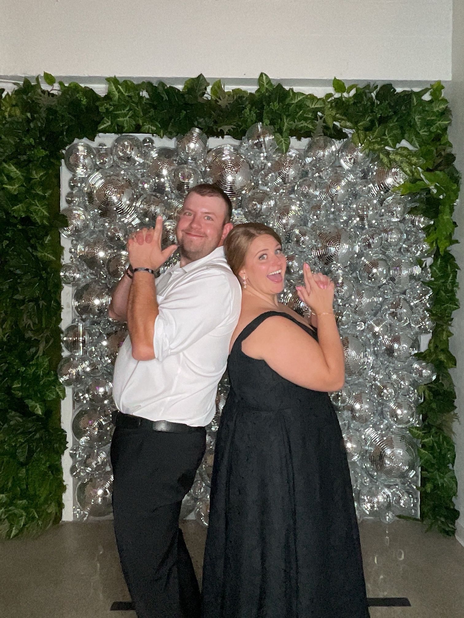 Two people pose back-to-back in front of a silver backdrop with green foliage