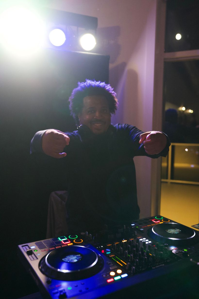 DJ at a mixing console with colorful lights, pointing toward the camera in a dimly lit room