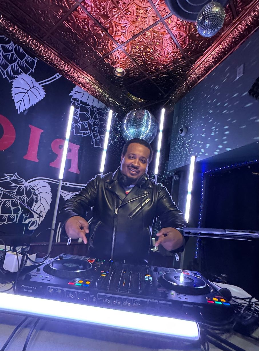 DJ smiling behind a mixer in a neon-lit booth with purple and blue lights