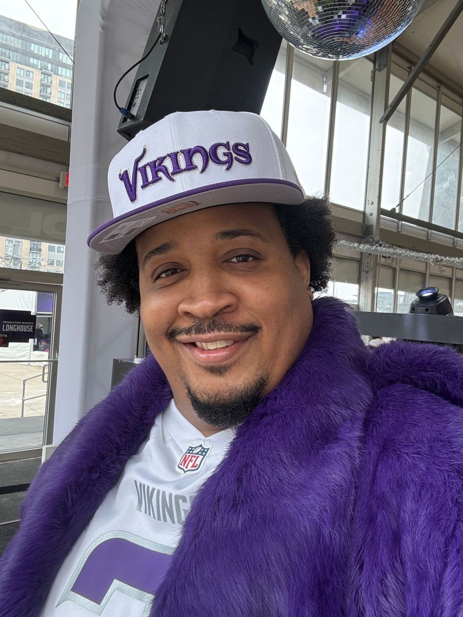Man smiling in a purple Vikings hat and jacket, standing indoors near a windowed area.