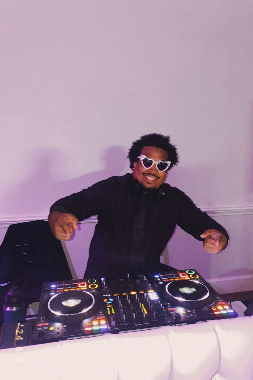 DJ in a black outfit and white sunglasses stands behind a lit mixing console, smiling and dancing.