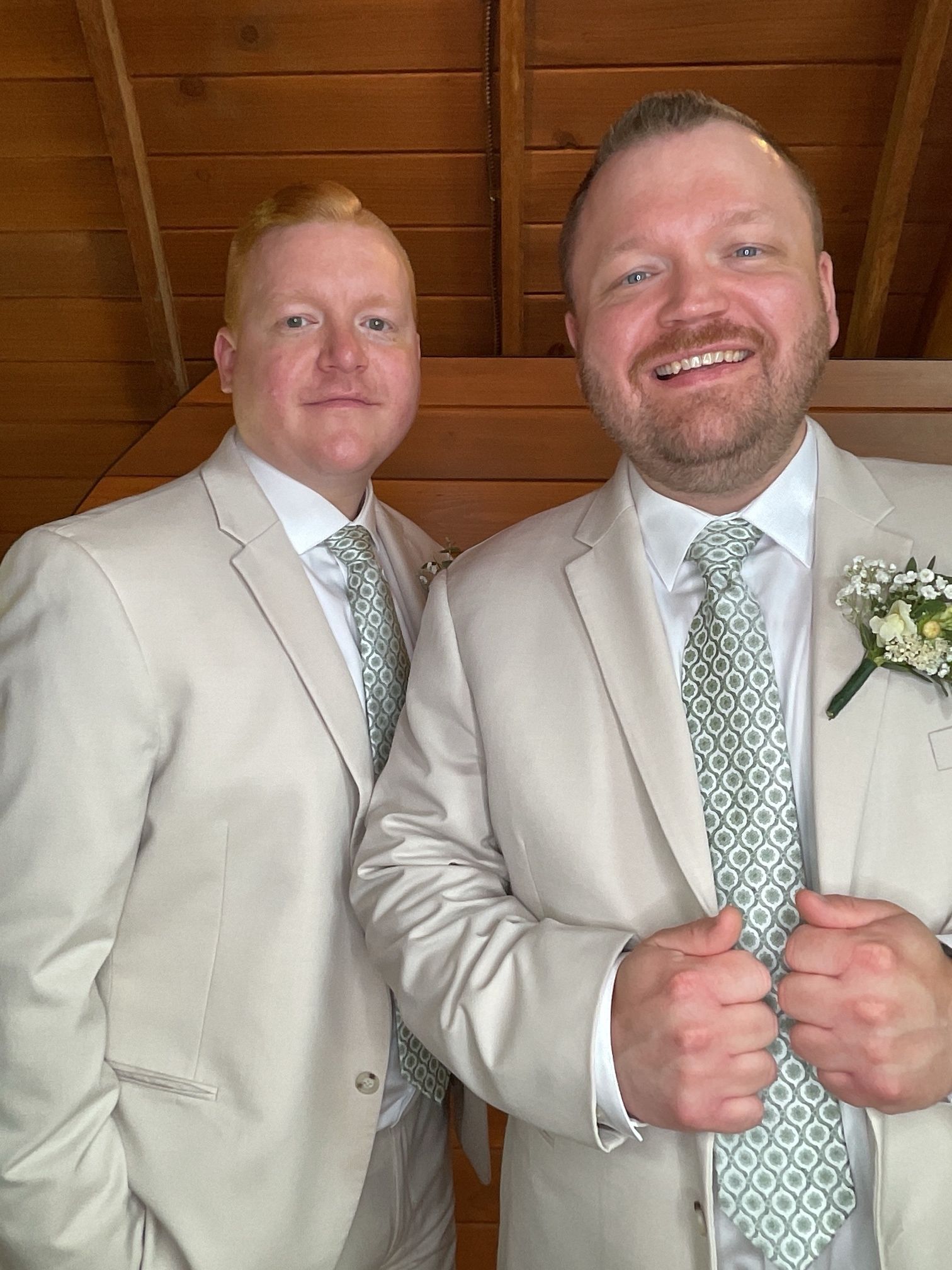 Two men in beige suits with mint green ties pose indoors, smiling in front of wood paneling.