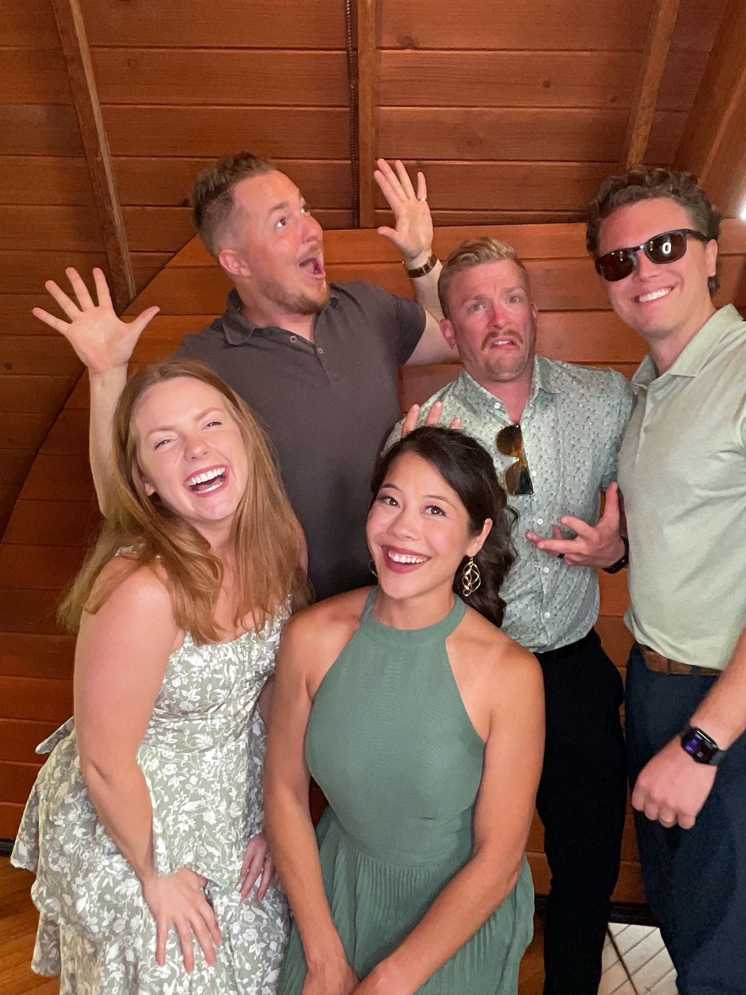 Five smiling people posing indoors against a wooden wall, with playful raised hands and casual outfits.