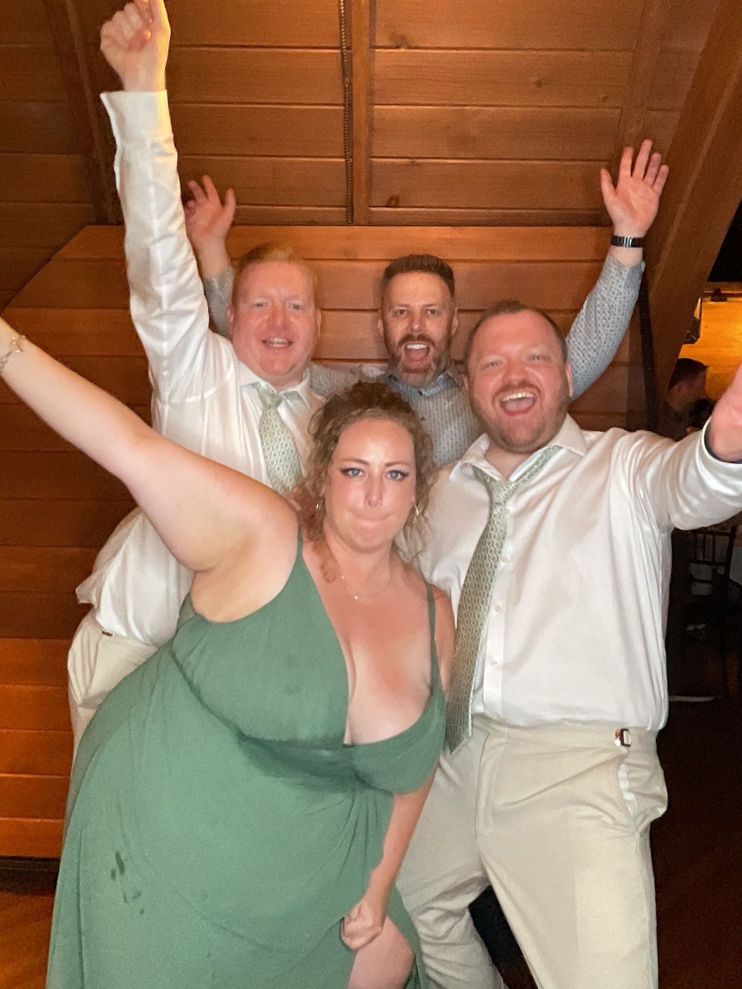 Four people pose indoors, smiling and raising their arms in front of wooden walls.