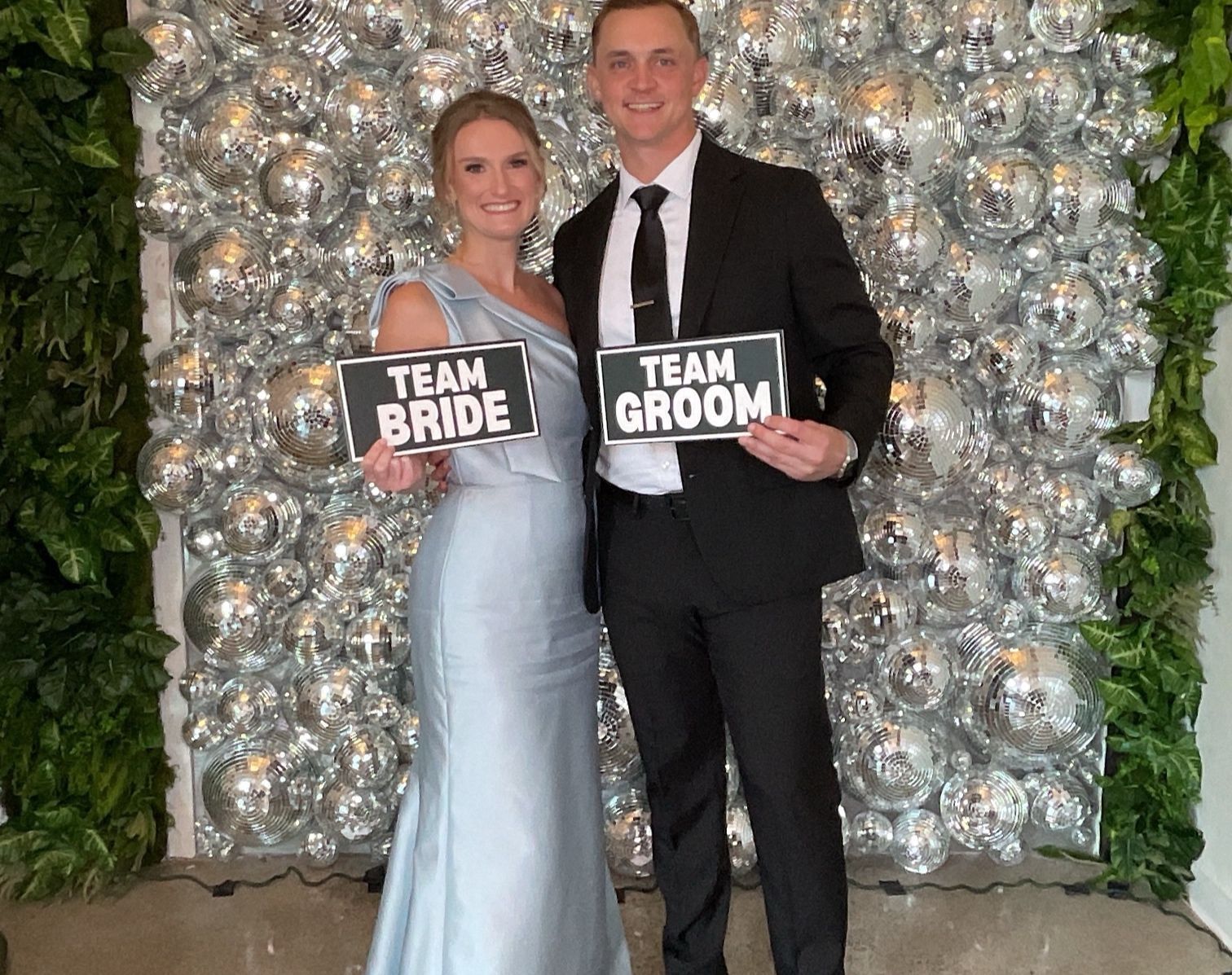 Bride and groom posing in formal wear, holding “Team Bride” and “Team Groom” signs in front of a silver backdrop