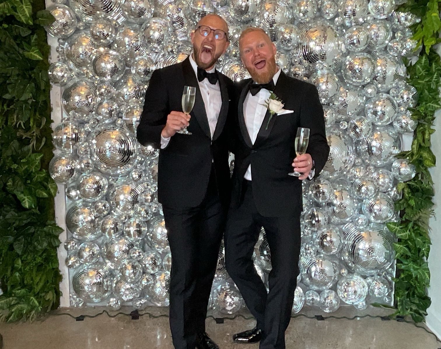 Two men in black tuxedos posing with champagne glasses in front of a silver balloon wall