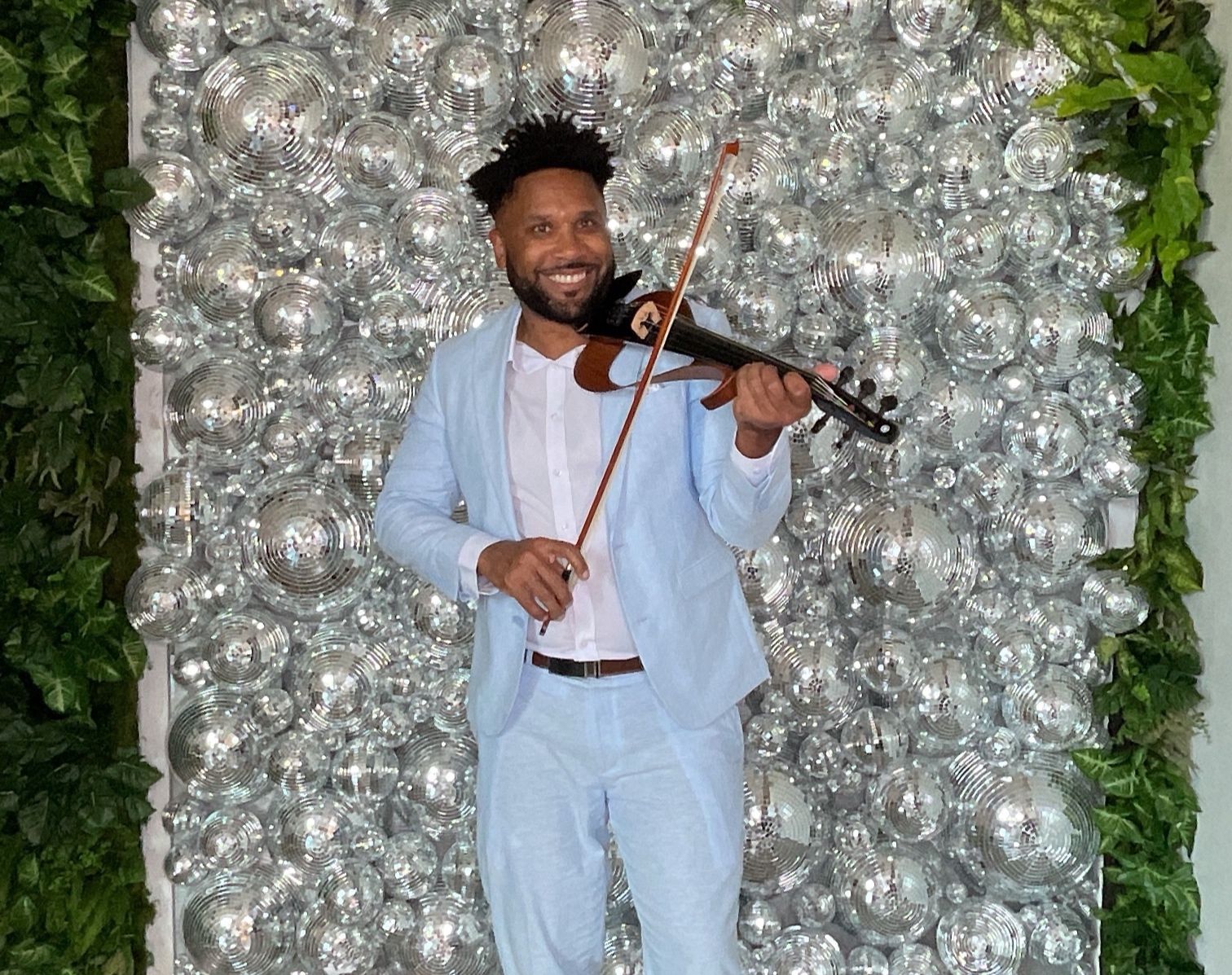 Man in a light blue suit smiling while playing violin in front of a silver floral wall