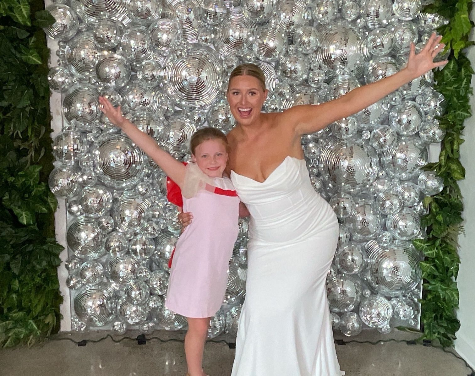 Two women in formal dresses pose with arms raised before a silver balloon wall.