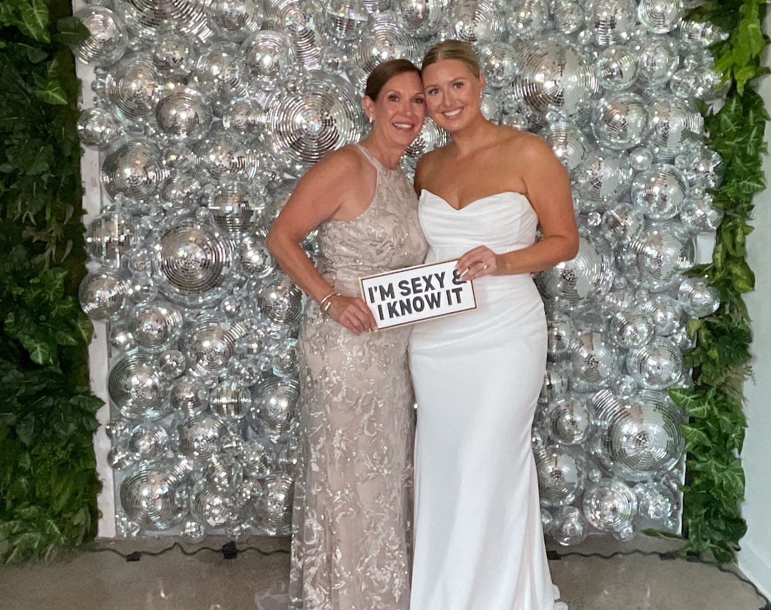 Two people posing in formal dresses before a silver balloon wall, holding a sign that says “Classy I know it.”
