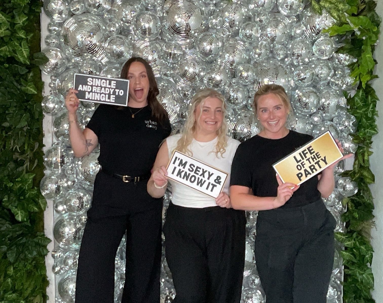 Three people posing before a silver balloon wall, holding playful signs, dressed in black and smiling.