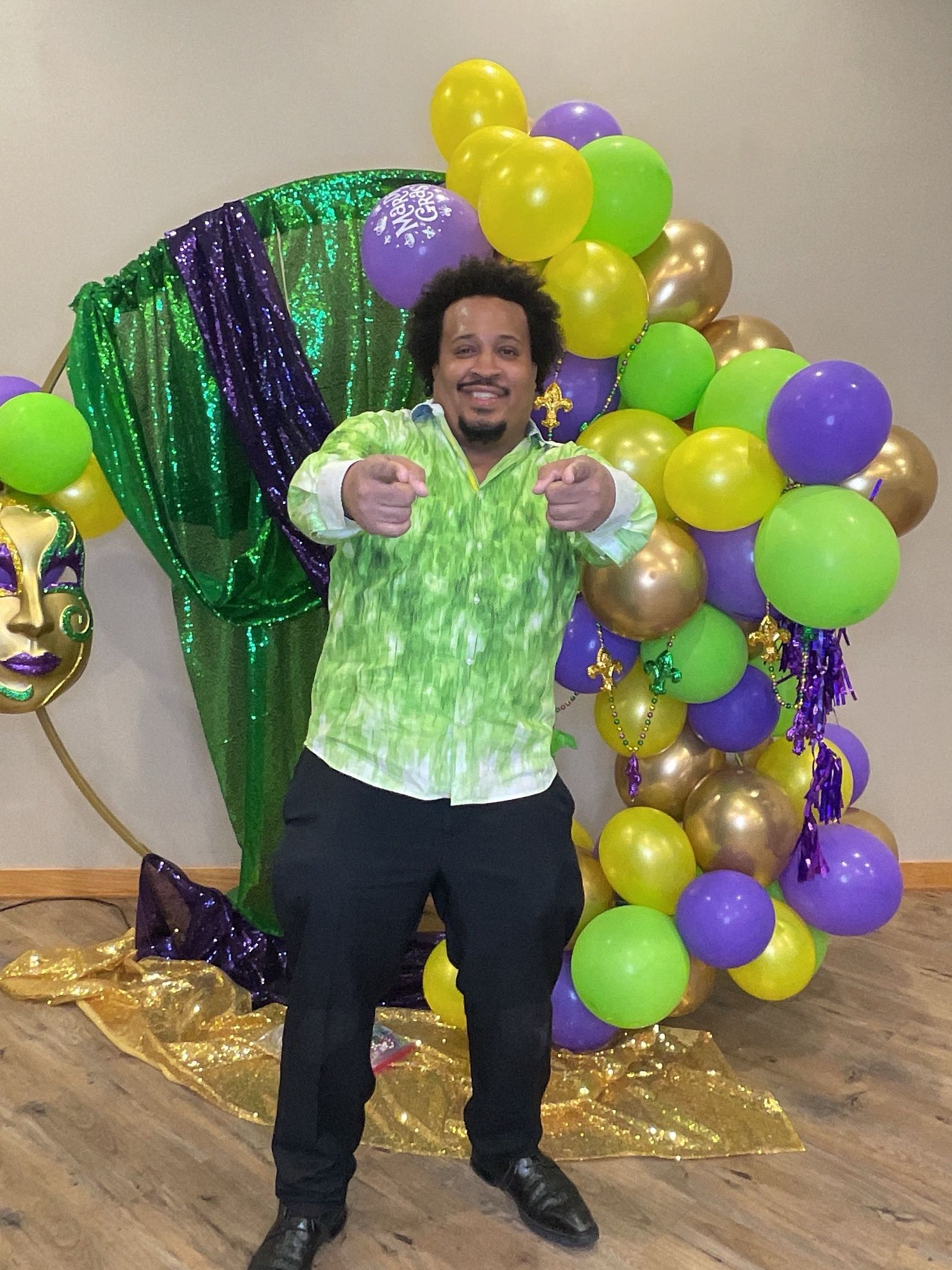 Smiling person in a green vest points at camera in front of green, yellow, and purple balloons.