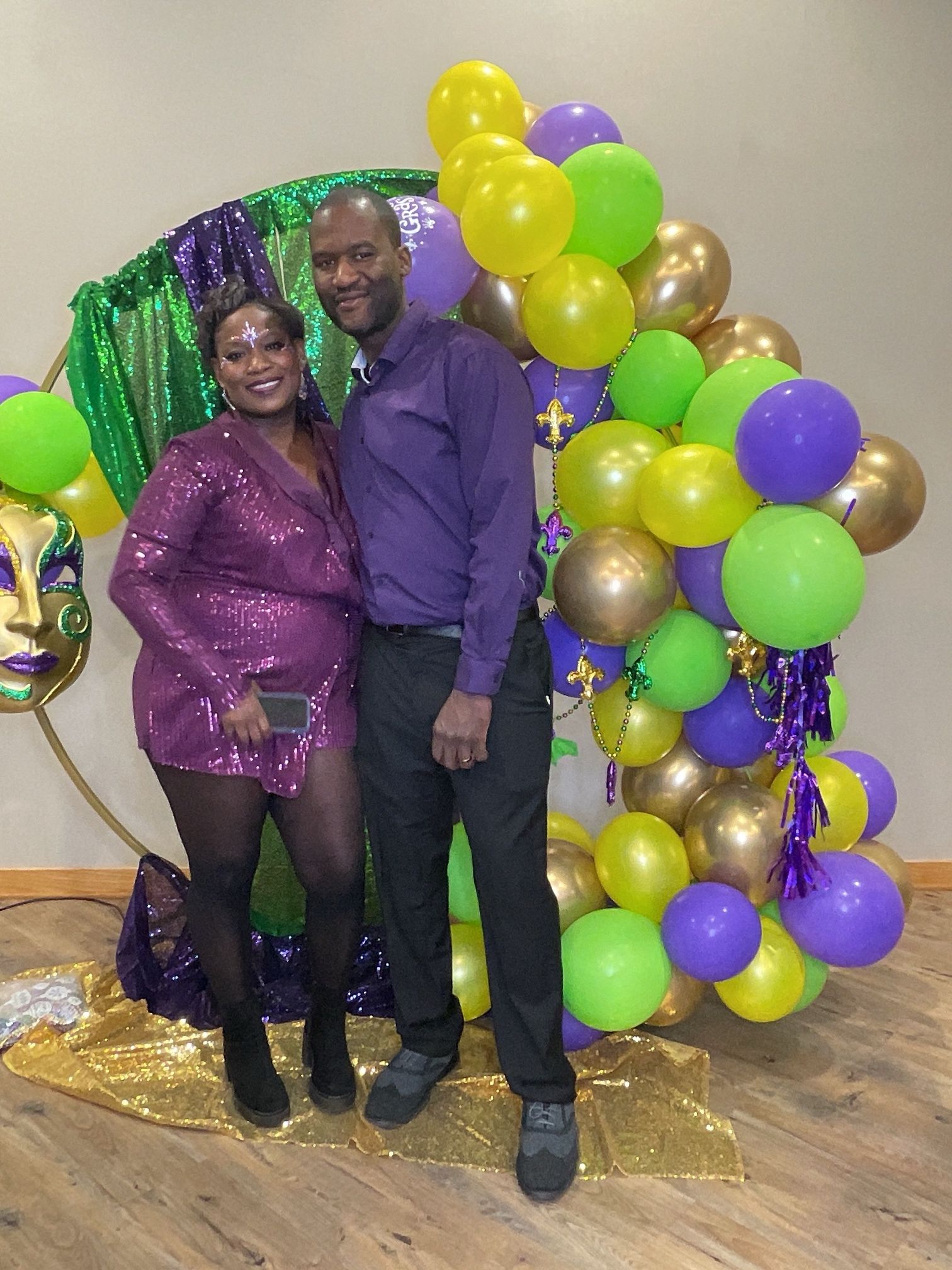 Two people posing by purple, gold, and green balloons at a festive party backdrop