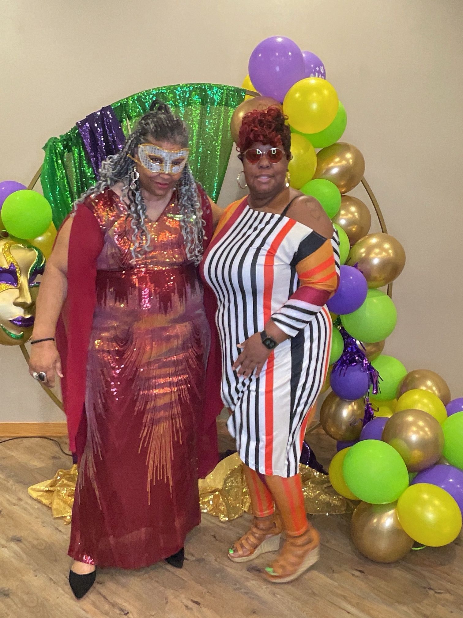 Two women in colorful dresses posing in front of green, purple, and gold balloon decorations.