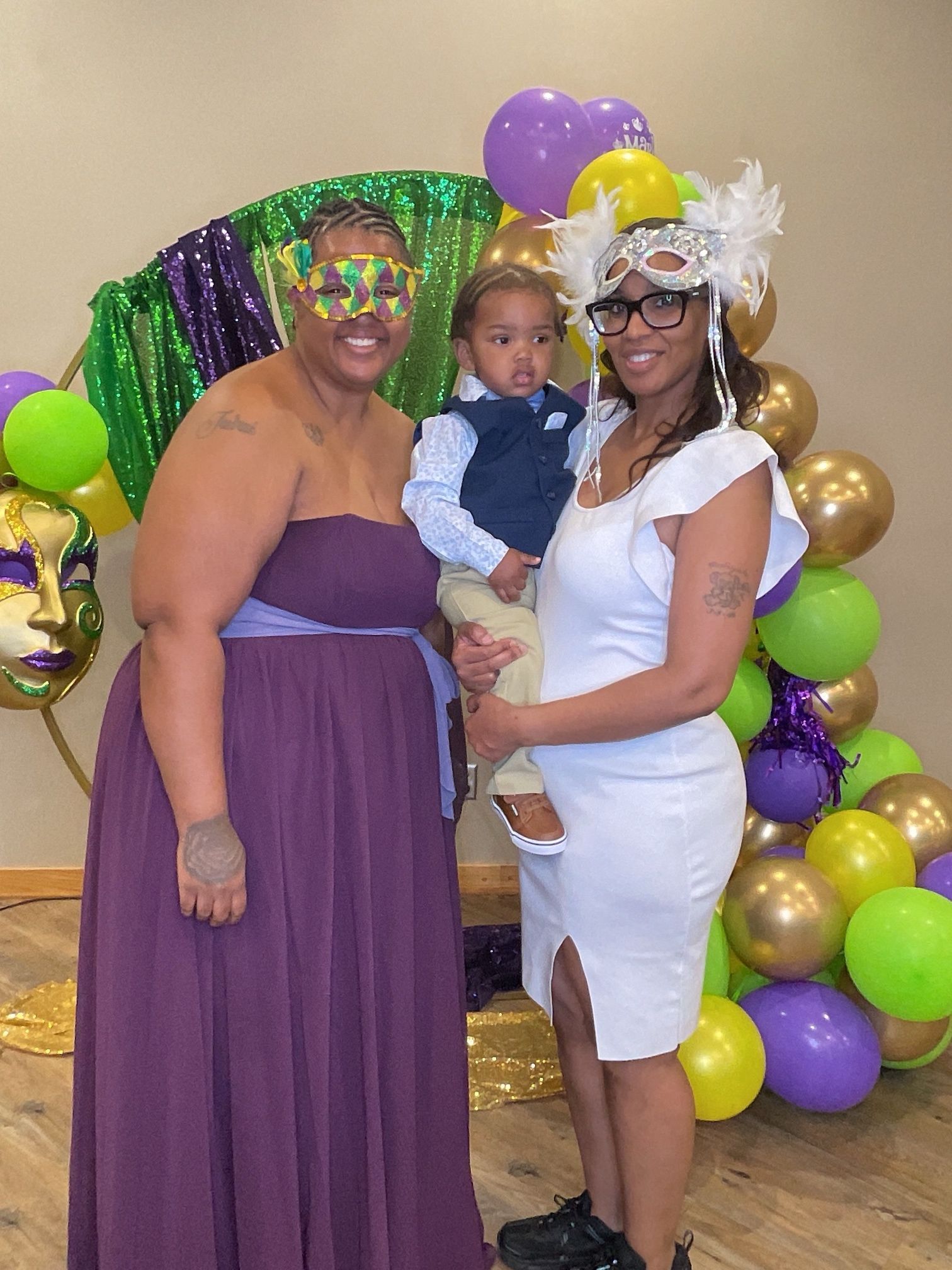 Three people posing at a balloon-decorated party, with purple, gold, and green decorations.