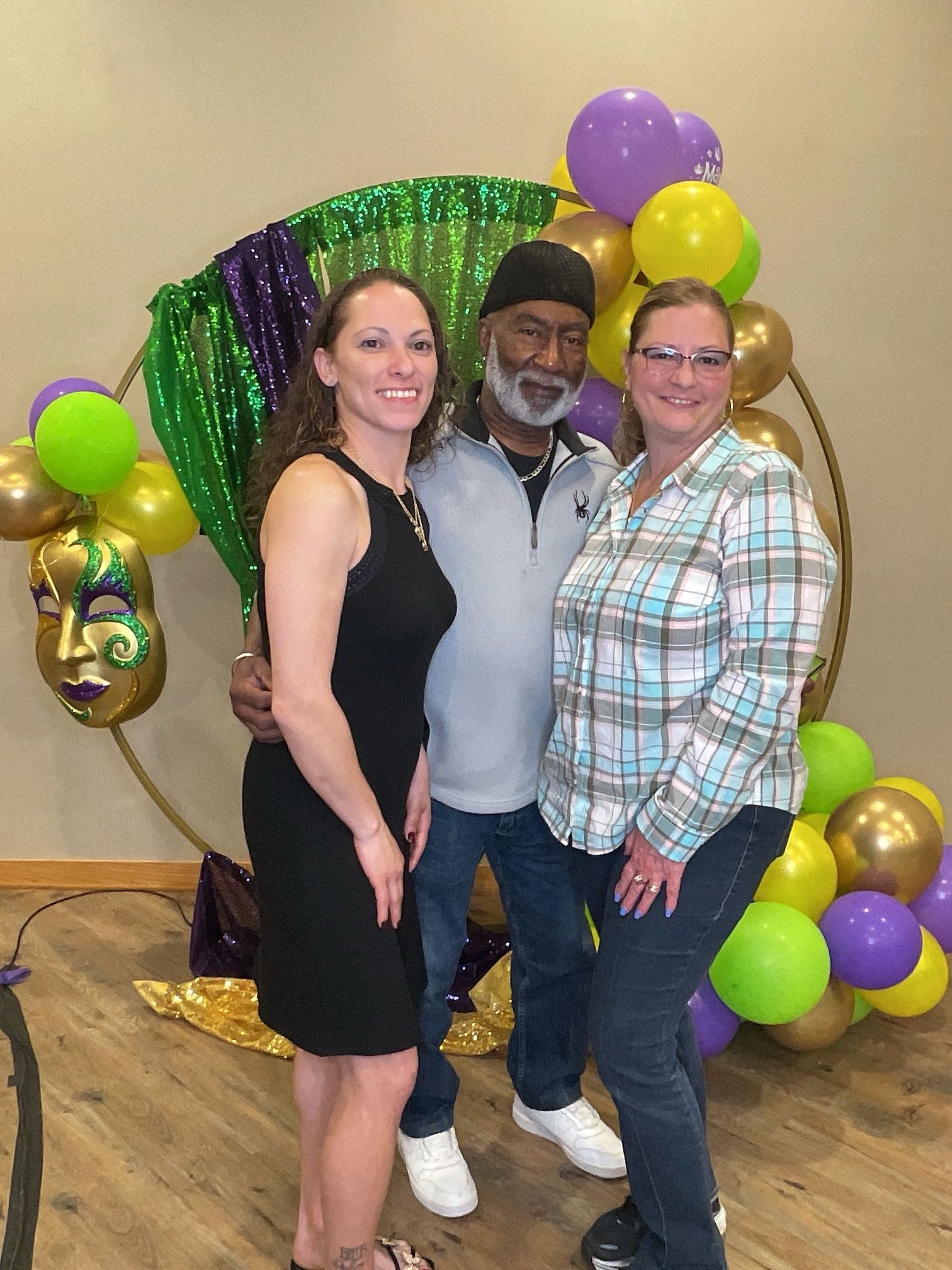 Three people posing before a gold, purple, and green balloon backdrop at a celebration.