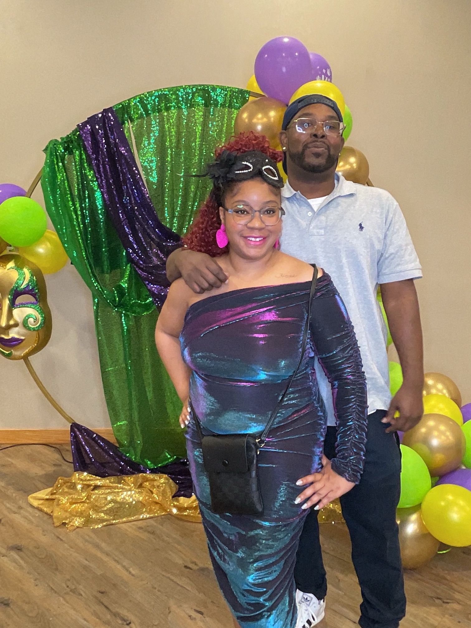 Two people posing in front of green and gold party decorations, one in a shimmering dress and one in a gray polo.