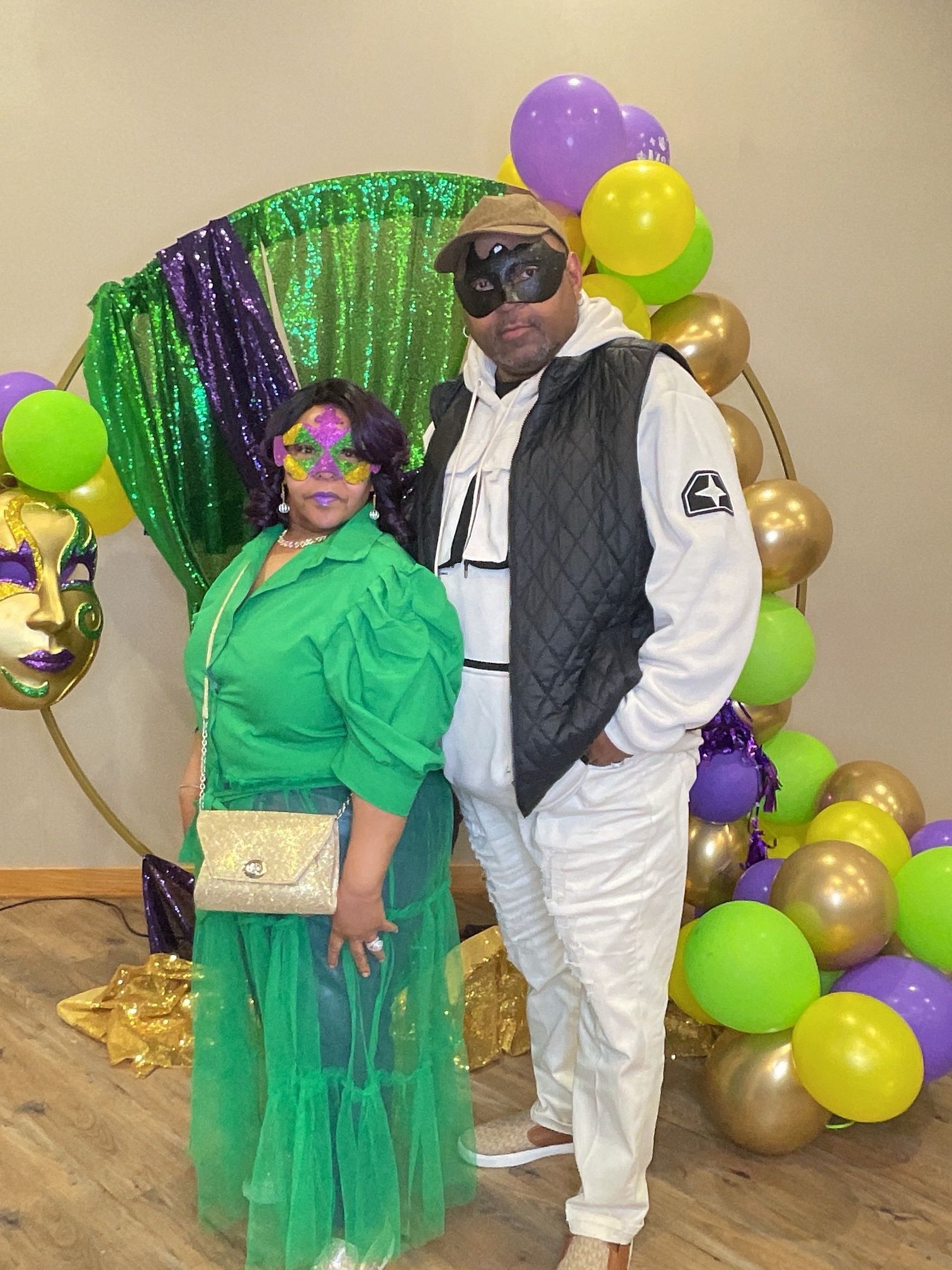 Two people posing in Mardi Gras costumes with green, purple, and gold decorations