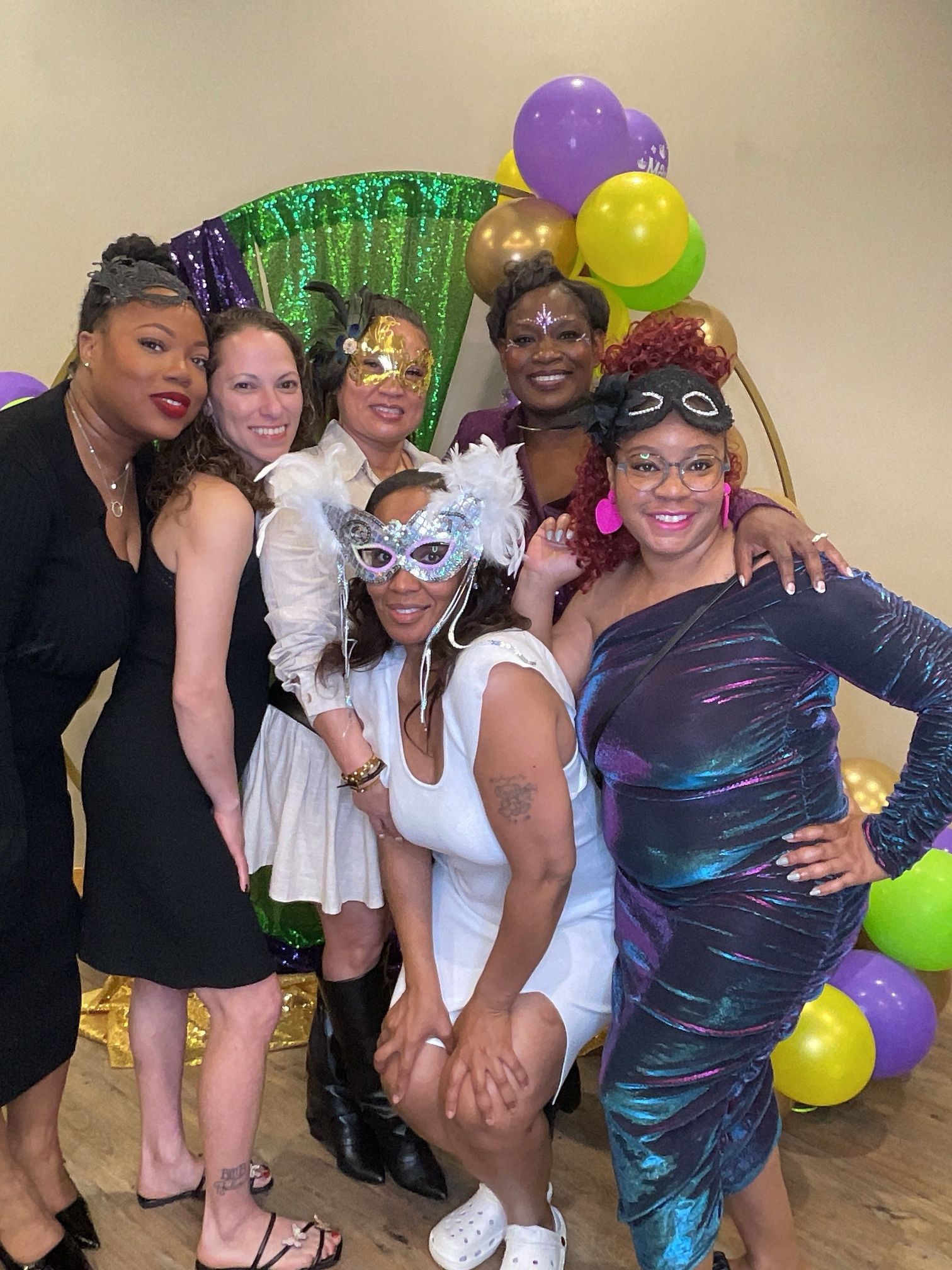 Five people posing in festive party attire with purple, gold, and green balloons and a glittery backdrop