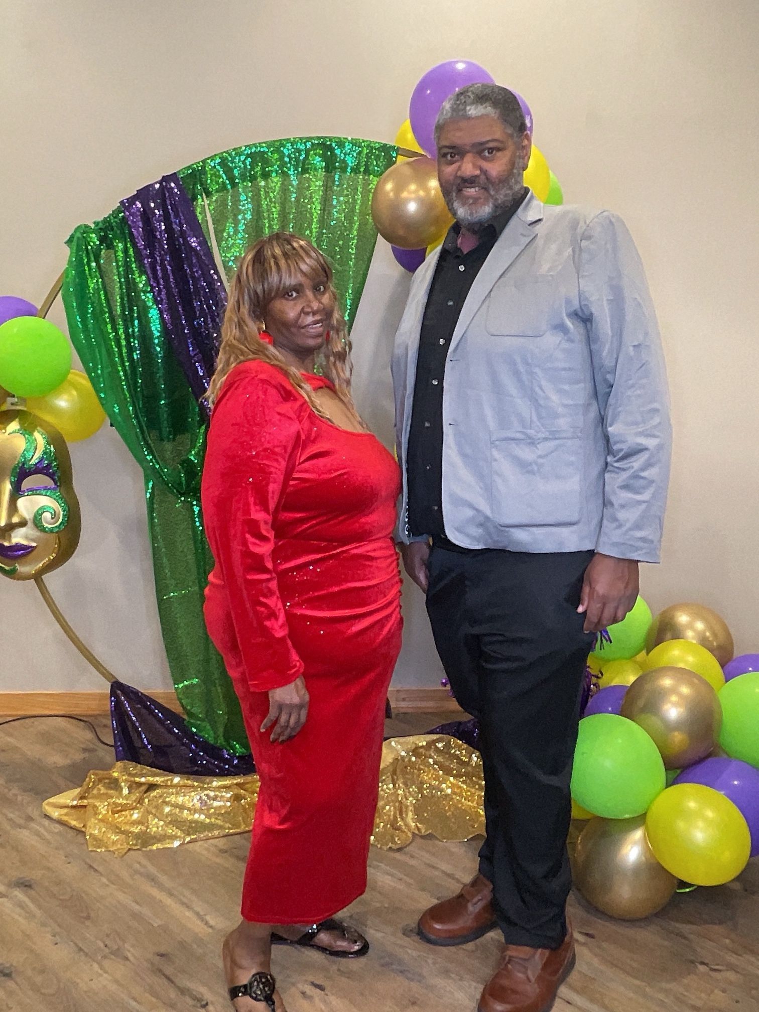 Two people posing at a colorful party backdrop with balloons and green, purple, and gold decorations.