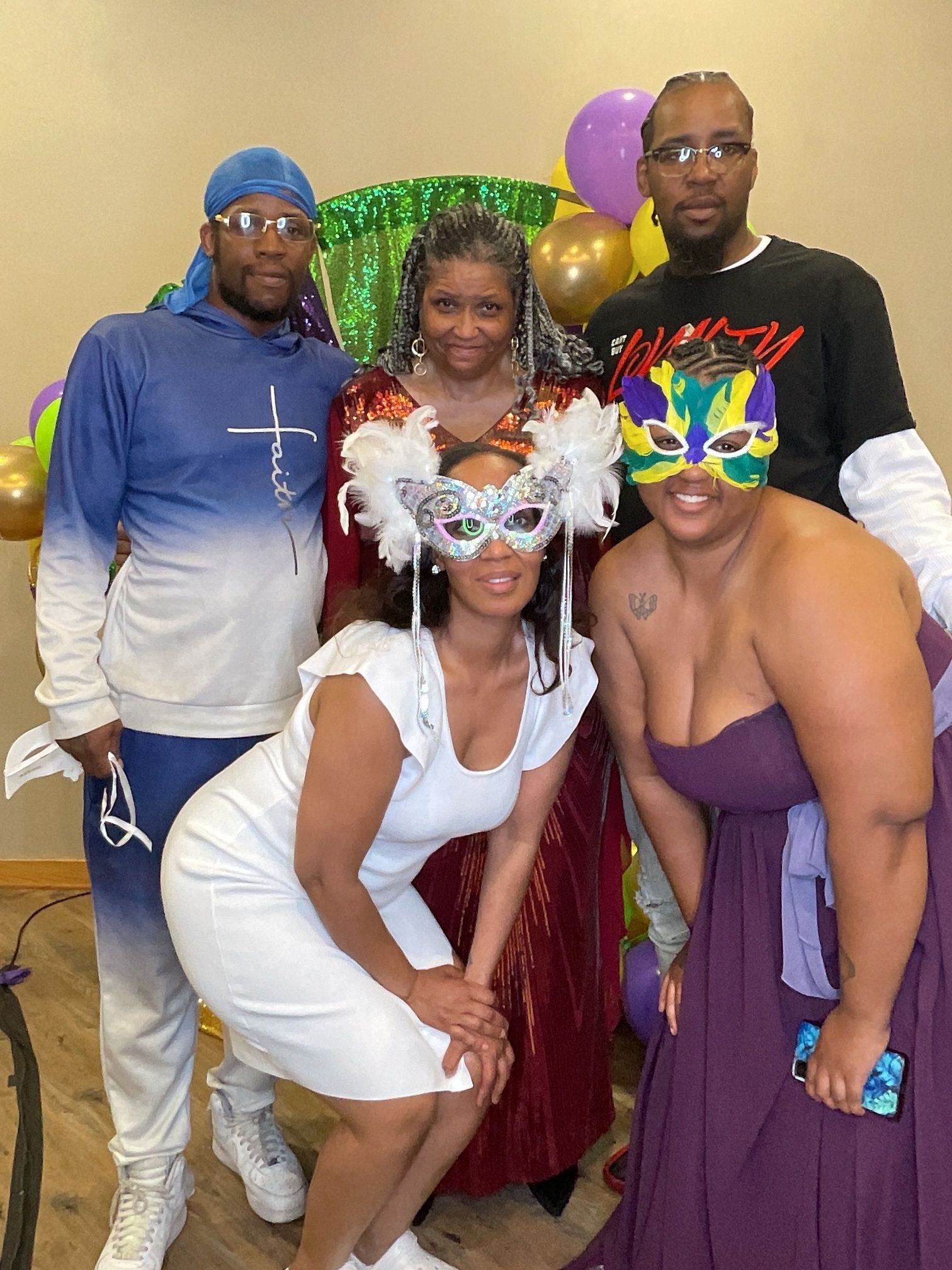 Four people posing in costumes and festive masks at a party with balloons and a shimmering backdrop.
