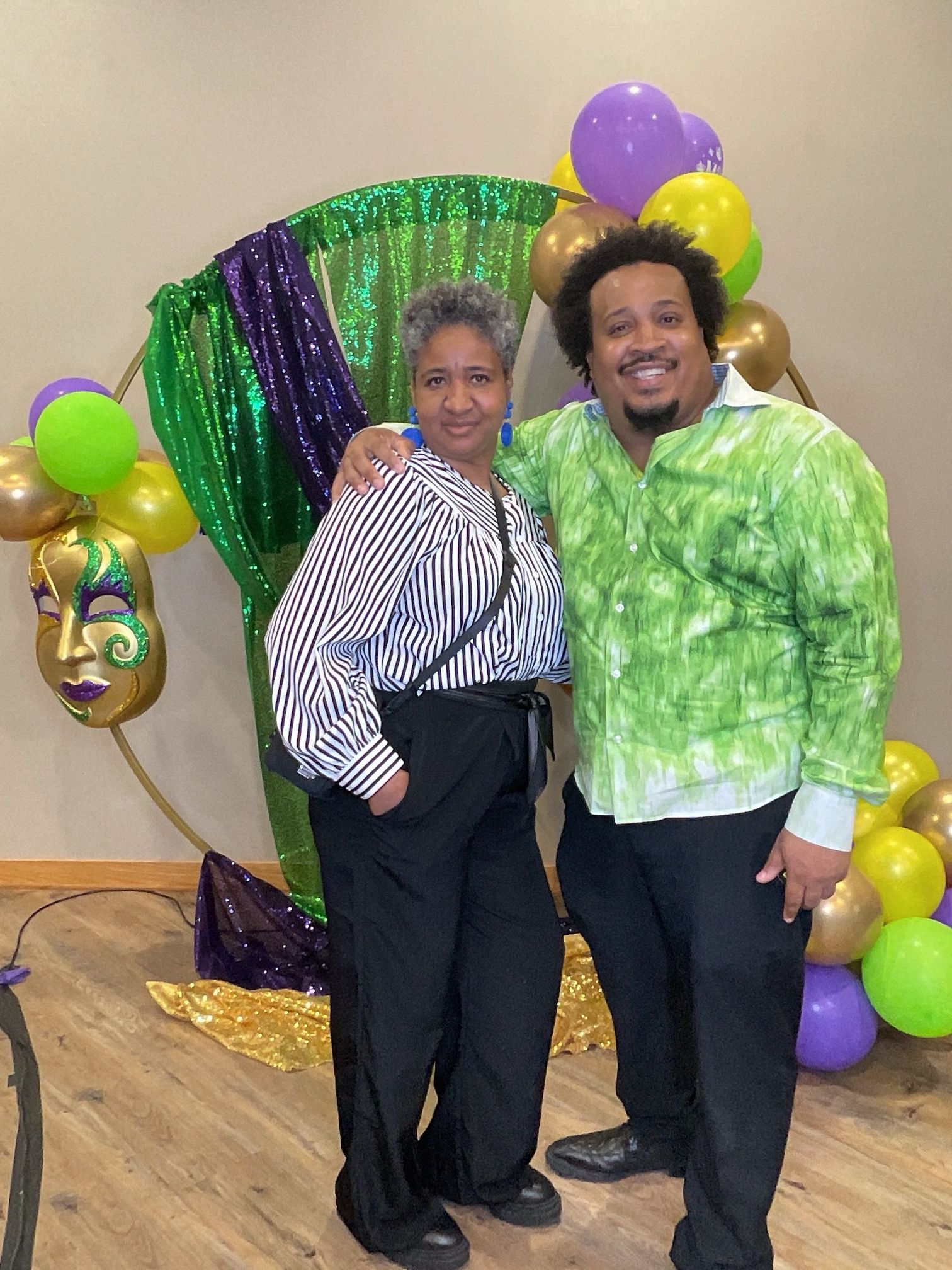 Two people posing together at a festive balloon backdrop with purple and green decorations.