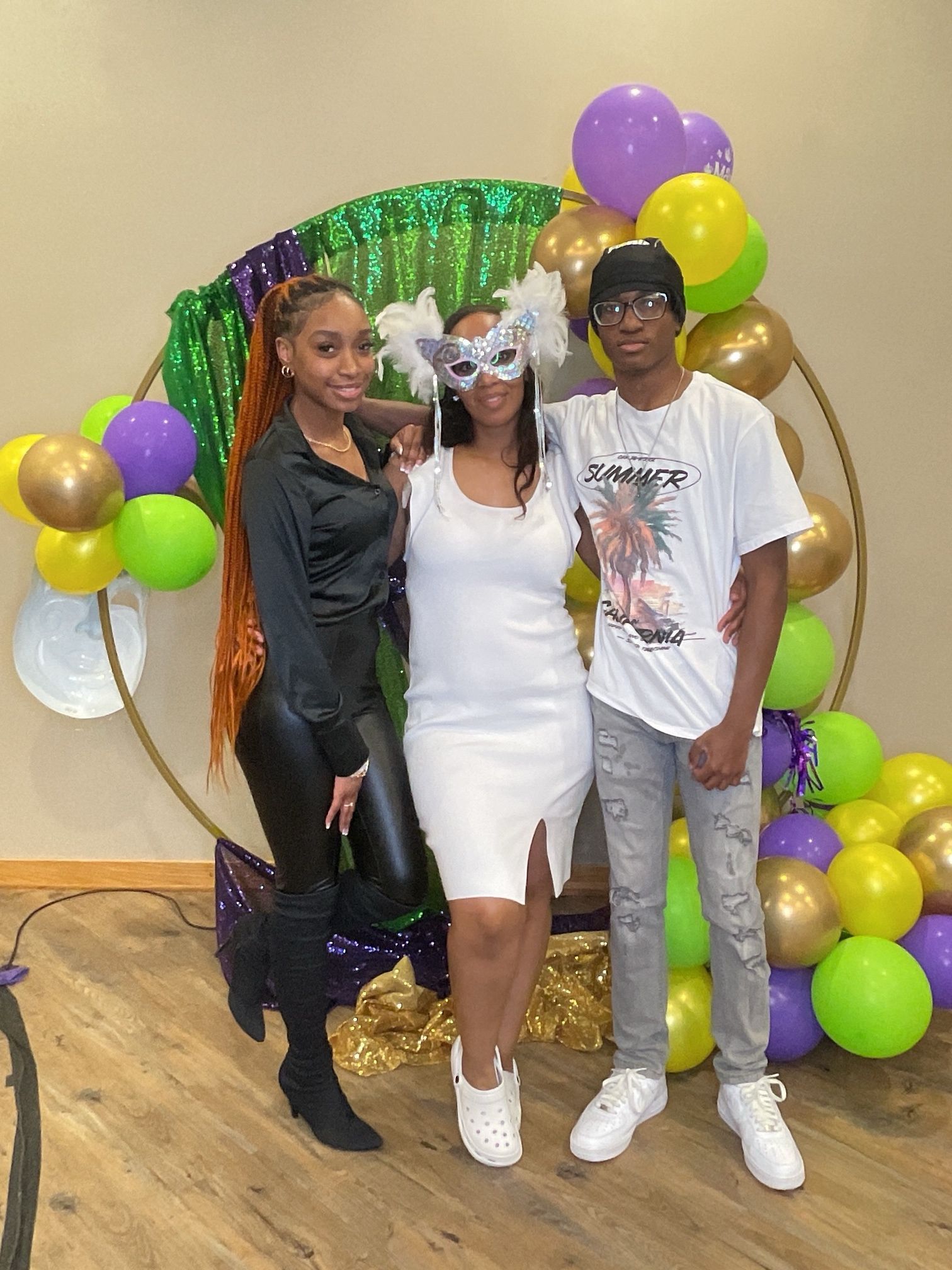 Three people posing in front of a green and gold balloon arch at a party.